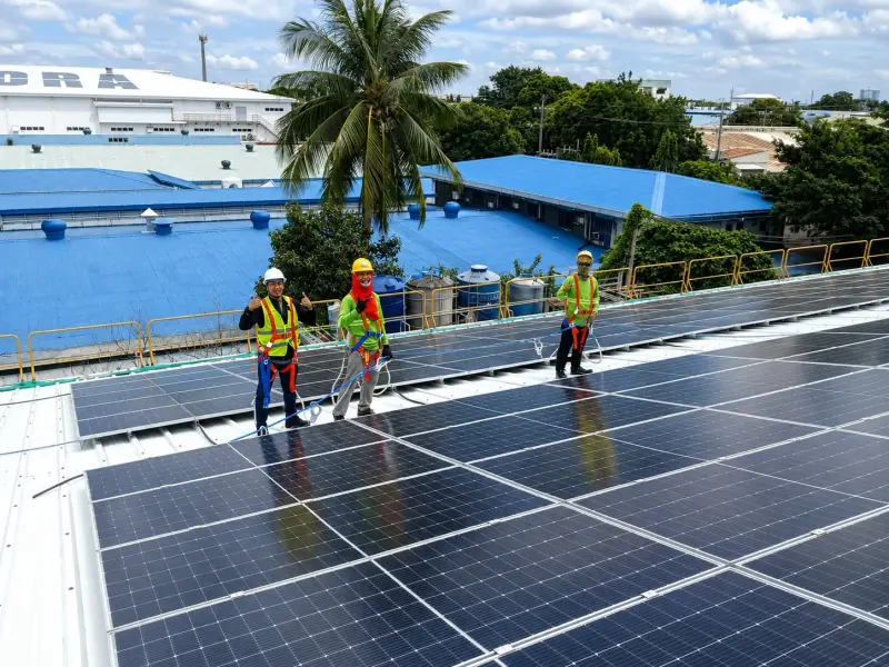 Three safety-equipped workers installing solar panels on an industrial rooftop with blue warehouses and equipment visible