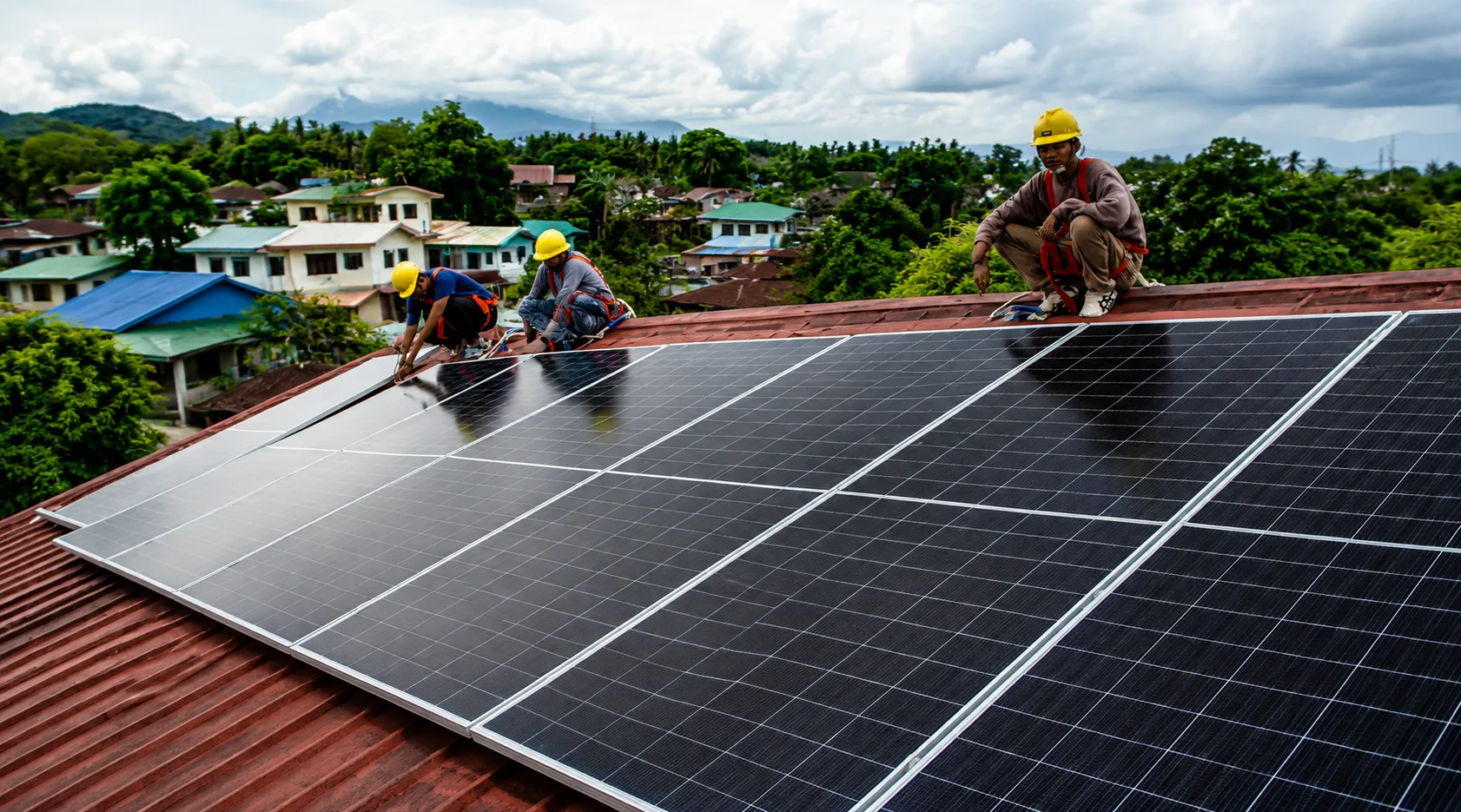 Three workers in safety helmets installing solar panels on a residential rooftop in a tropical village setting