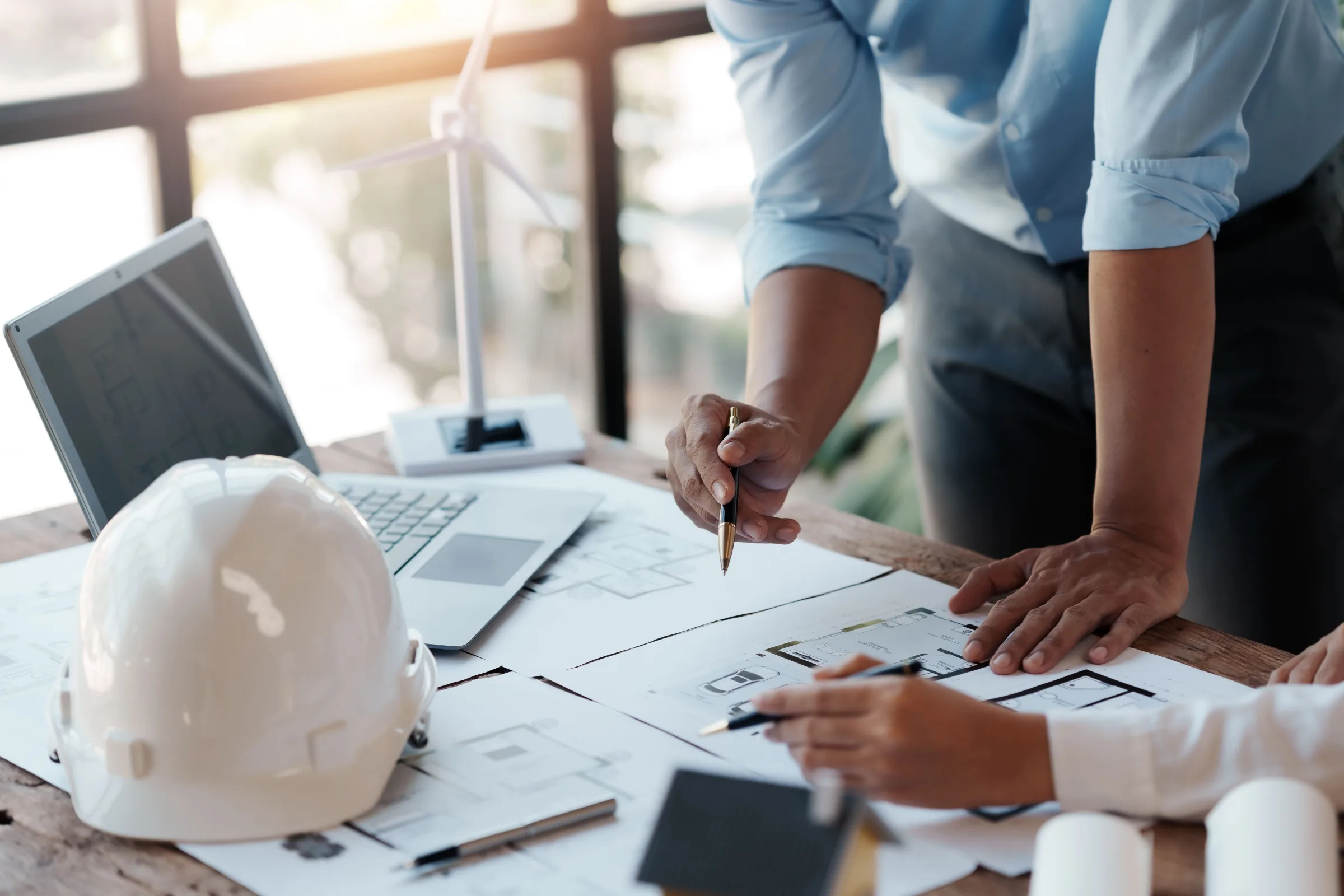 Architects and engineers reviewing building blueprints and plans at a desk with a hard hat and laptop