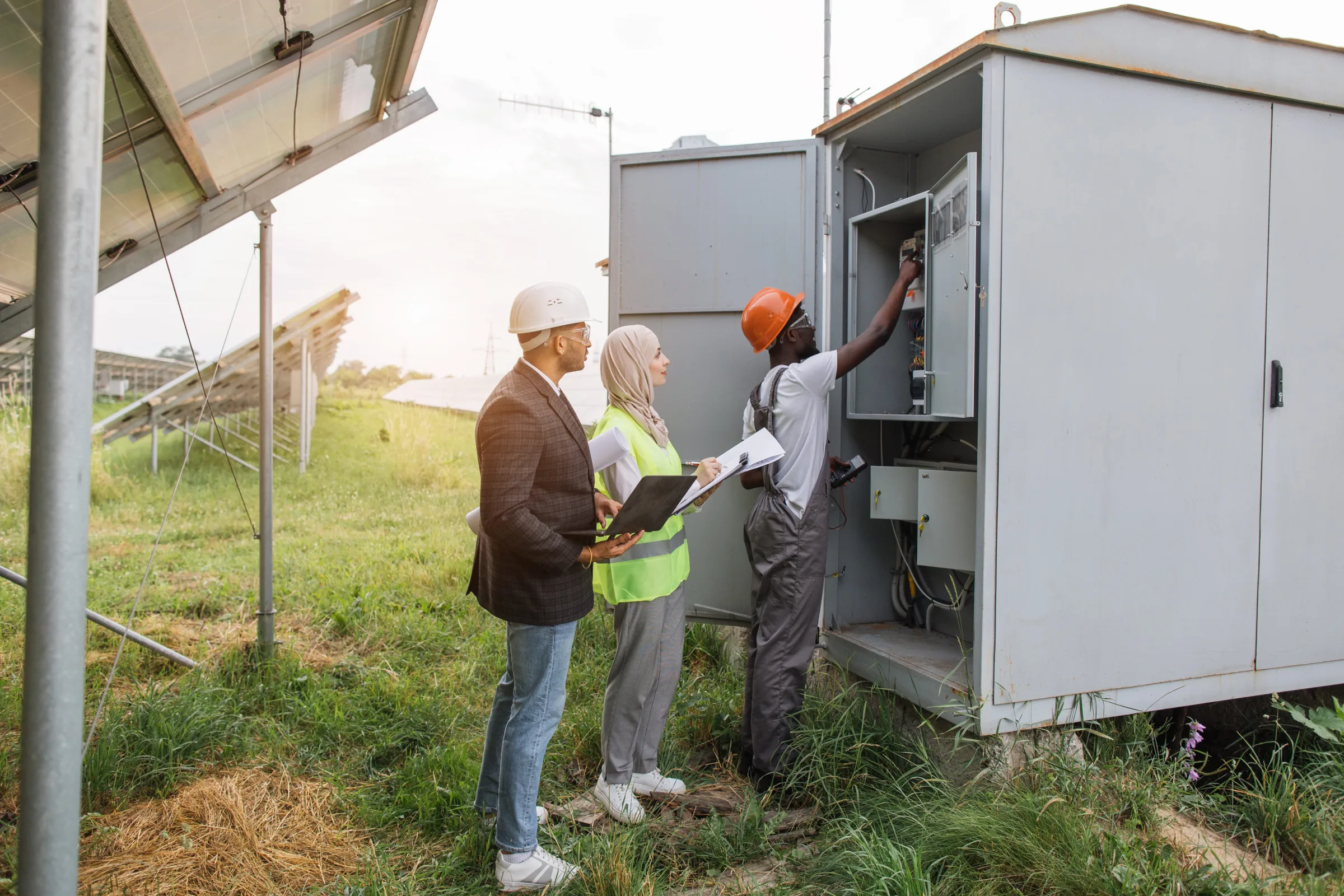 Three professionals inspecting electrical equipment at a solar panel installation site